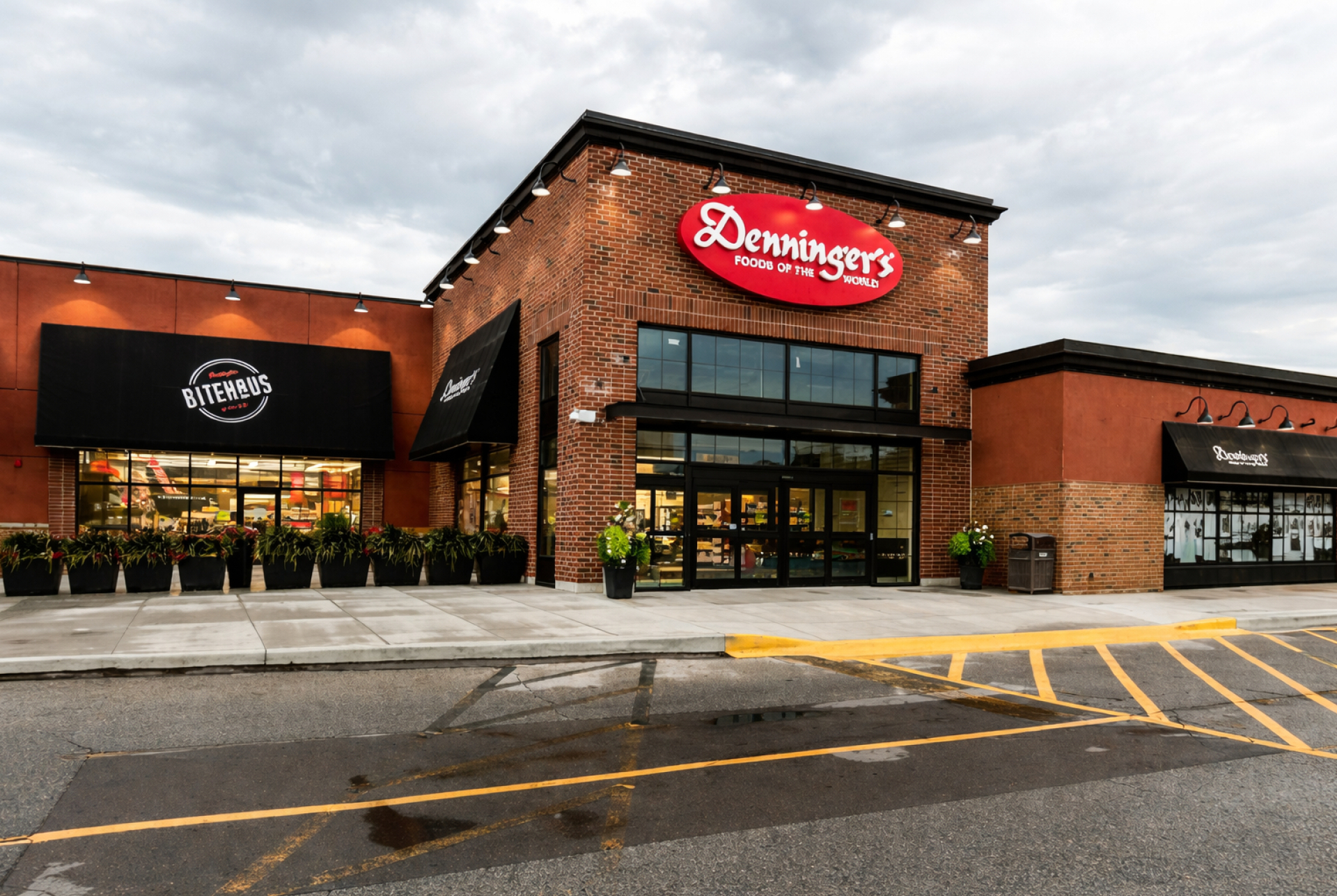 A Denninger’s grocery store exterior with a brick facade and a red “Denninger’s Foods of the World” sign above the main entrance. Large glass doors, storefront windows, and planters line the sidewalk under an overcast sky.